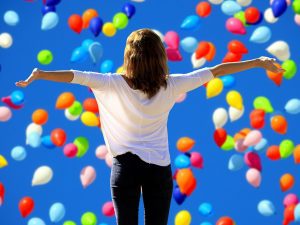 A lady has arms out looking up with joy amongst colourful ballons